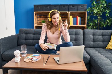 Young beautiful woman practicing painting looking at online tutorial surprised with an idea or question pointing finger with happy face, number one 
