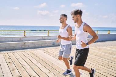 İki İspanyol erkek, sportif, gülümseyen bir çift, deniz kenarında koşuyorlar.