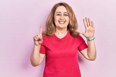 Orta yaşlı beyaz kadın, günlük kıyafetler giyip altı numaralı parmaklarıyla işaret ederken kendinden emin ve mutlu bir şekilde gülümsüyor.. 