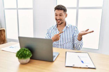 Young handsome man with beard working at the office using computer laptop amazed and smiling to the camera while presenting with hand and pointing with finger. 