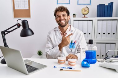 Young hispanic dentist man working at medical clinic cheerful with a smile of face pointing with hand and finger up to the side with happy and natural expression on face 