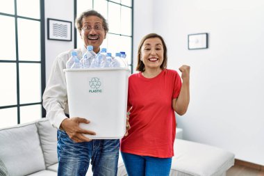 Middle age interracial couple holding recycling bin with plastic bottles at home screaming proud, celebrating victory and success very excited with raised arms 