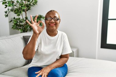 Olgun İspanyol kadın evdeki kanepede oturmuş olumlu gülümsüyor ve el ve parmaklarıyla 