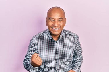 Orta yaşlı, gündelik kıyafetler giyen, mutlu ve heyecanlı bir Latin adam kolları havada, gülümseyen ve başarı için bağıran, kazanan bir jest yapıyor. kutlama konsepti. 