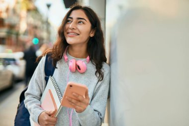 Genç ortadoğulu kız şehirde akıllı telefon kullanarak mutlu bir şekilde gülümsüyor..