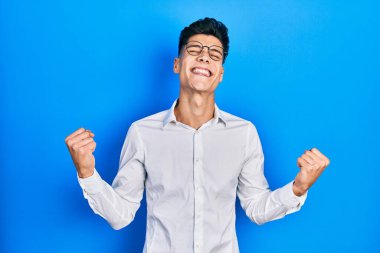 Gündelik kıyafetler ve gözlük takan genç İspanyol adam mutlu ve heyecanlı bir şekilde kolları havada, gülümseyen ve başarı için bağıran kazanan bir jest yapıyor. kutlama konsepti. 