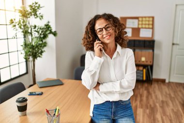 Orta yaşlı İspanyol kadın gülümsüyor ofisteki akıllı telefondan konuşuyor.