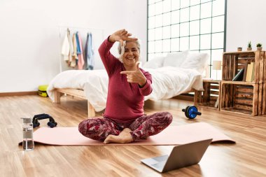 Evde gri saç eğitimi almış orta yaşlı bir kadın laptopta egzersiz videosu seyrediyor gülümsüyor ve elleri ve parmakları mutlu yüzüyle çerçeve yapıyor. yaratıcılık ve fotoğrafçılık kavramı. 