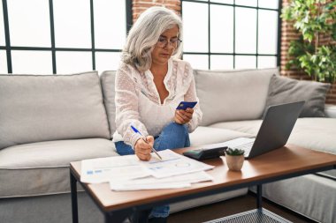 Evde dizüstü bilgisayar ve kredi kartı kullanan orta yaşlı kadın.
