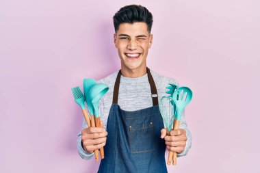 Profesyonel fırıncı önlüğü giyen genç İspanyol adam yemek aletleri tutuyor kameraya seksi bir ifadeyle bakıyor, neşeli ve mutlu bir yüzle.. 