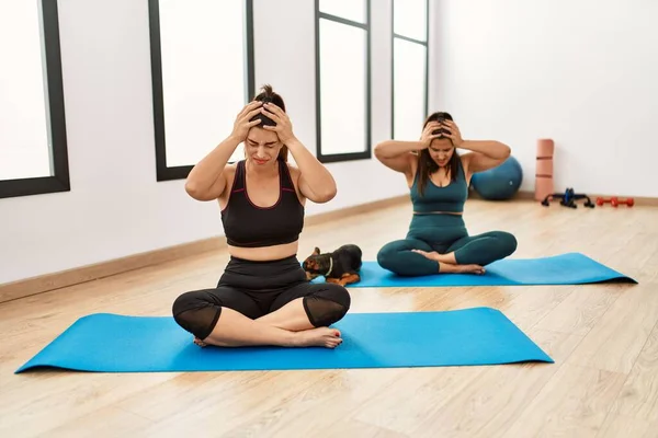 Two hispanic women friends training at the gym sitting on yoga mat with dog suffering from headache desperate and stressed because pain and migraine. hands on head.