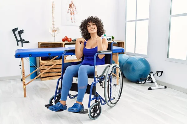 Young middle eastern woman sitting on wheelchair at physiotherapy clinic looking confident with smile on face, pointing oneself with fingers proud and happy.