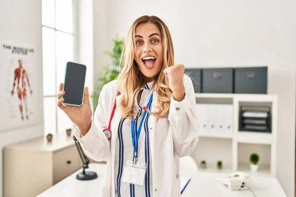 Young Blonde Doctor Woman Working Clinic Showing Smartphone Screen Screaming — Stock Photo, Image