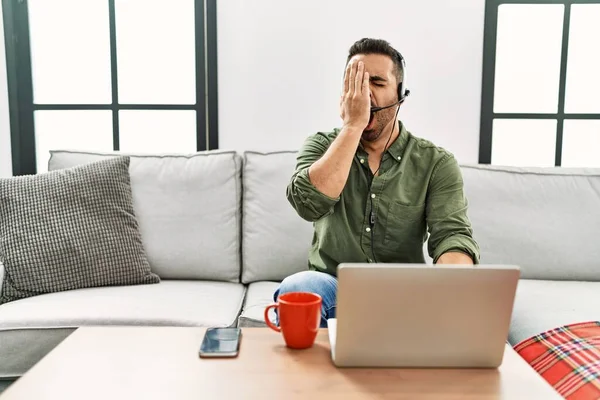 Young hispanic man with beard wearing call center agent headset working from home yawning tired covering half face, eye and mouth with hand. face hurts in pain.