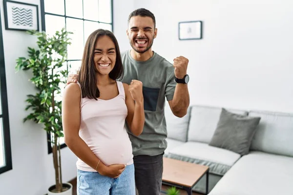 Young interracial couple expecting a baby, touching pregnant belly very happy and excited doing winner gesture with arms raised, smiling and screaming for success. celebration concept.