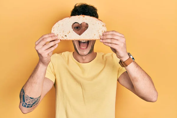 Young hispanic man holding bread with heart shape over eye celebrating crazy and amazed for success with open eyes screaming excited.