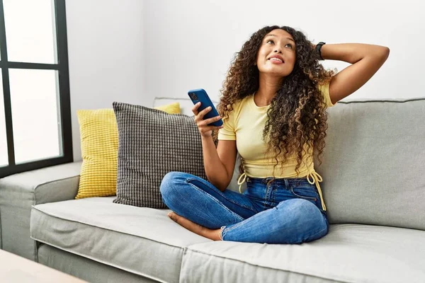 自宅でソファに座っている美しいヒスパニック系の女性は スマートフォンを使用して自信を持って手をジェスチャーで髪に触れ笑顔 魅力的でファッショナブルなポーズ — ストック写真