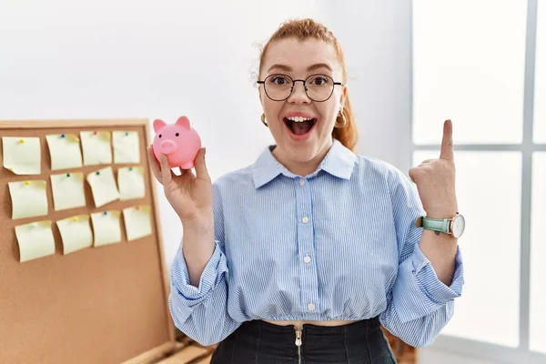 Joven Pelirroja Sosteniendo Alcancía Oficina Sorprendida Con Una Idea Pregunta — Foto de Stock