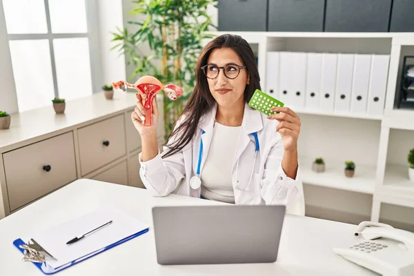 Young hispanic doctor woman holding anatomical female genital organ and birth control pills smiling looking to the side and staring away thinking.