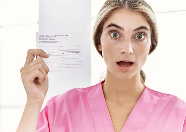 Young blonde woman holding covid record card scared and amazed with open mouth for surprise, disbelief face