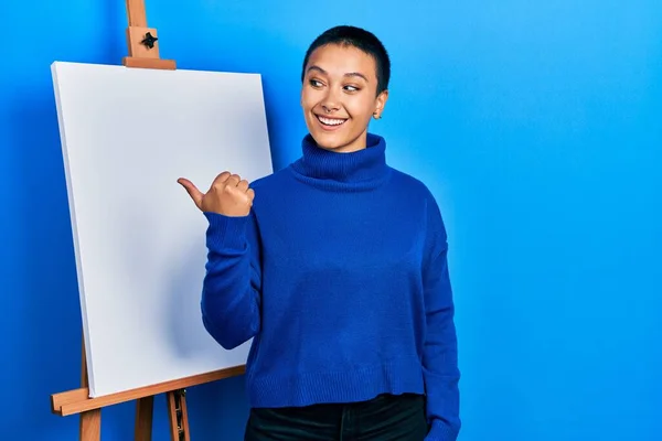 Beautiful hispanic woman with short hair standing by painter easel stand smiling with happy face looking and pointing to the side with thumb up.
