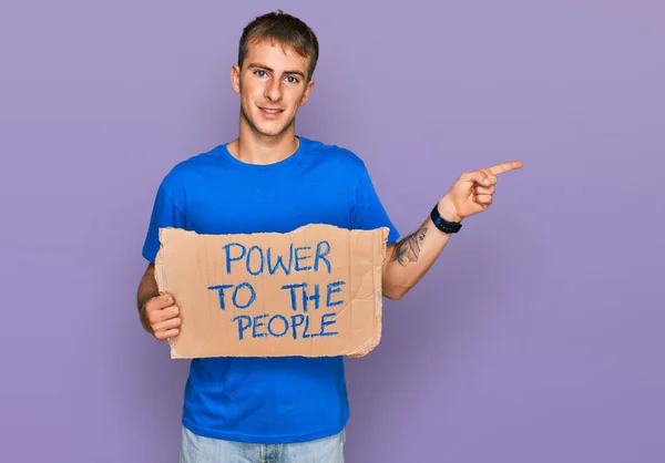 Young blond man holding power to the people banner smiling happy pointing with hand and finger to the side