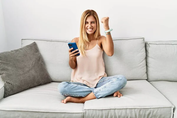 Blonde beautiful young woman sitting on the sofa at home using smartphone angry and mad raising fist frustrated and furious while shouting with anger. rage and aggressive concept.