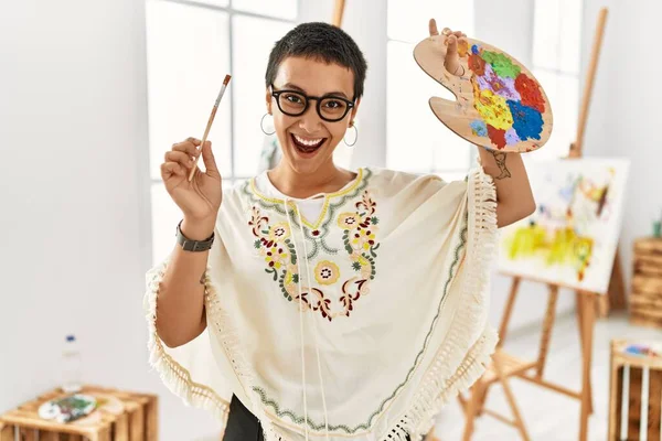 Resim stüdyosunda elinde fırça ve palet tutarak gülümseyen genç İspanyol kadın.