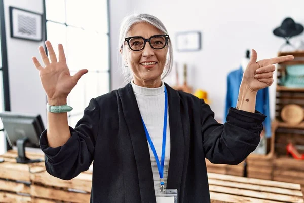 Middle age grey-haired woman working as manager at retail boutique showing and pointing up with fingers number seven while smiling confident and happy.