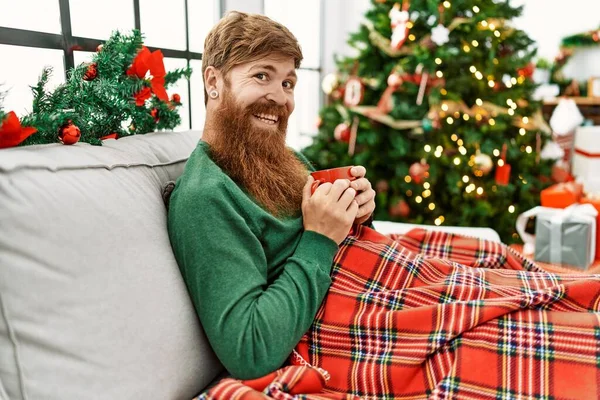 Young redhead man drinking coffee sitting by christmas tree at home