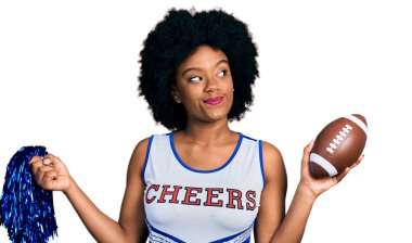 Young african american woman wearing cheerleader uniform holding pompom and football ball smiling looking to the side and staring away thinking. 