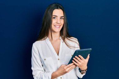 Young brunette teenager using touchpad device looking positive and happy standing and smiling with a confident smile showing teeth 