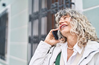 Orta yaşlı bir kadın gülümsüyor. Sokakta akıllı telefondan konuşuyor.