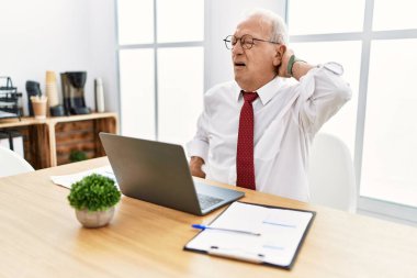 Ofiste çalışan kıdemli bir adam bilgisayar kullanıyor. Boyun ağrısı çekiyor, boynuna elle dokunuyor, kas ağrısı çekiyor. 