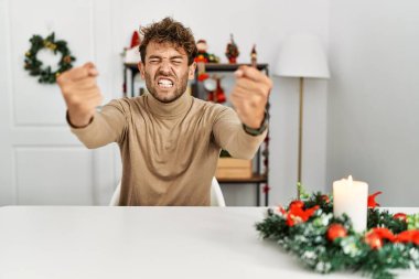 Noel süslemesi sırasında masanın üzerinde oturan sakallı yakışıklı genç adam kızgın ve kızgın yumruklarını kaldırırken sinirli ve öfkeliydi. öfke ve agresif konsept. 