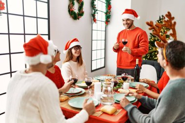 Mutlu bir şekilde gülümseyen bir grup genç Noel yemeği yiyor ve evde kadeh kaldırıyor..