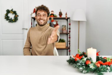 Noel süslemesi masasında oturan sakallı yakışıklı genç adam selamlaşma ve selamlama olarak gülümseyerek tokalaşıyor. başarılı iş. 