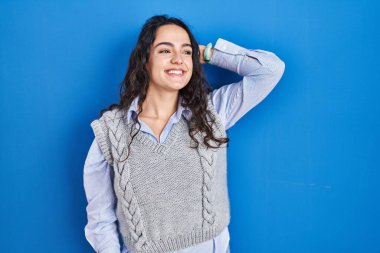 Genç esmer kadın mavi arka planda duruyor. Kendine güvenen, el hareketleriyle saçlara dokunan, çekici ve modaya uygun poz veren. 