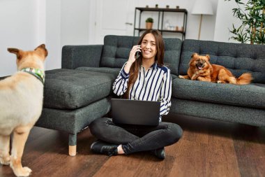 Genç İspanyol kadın dizüstü bilgisayar kullanıyor ve akıllı telefonla konuşuyor evde köpeklerle yerde oturuyor.
