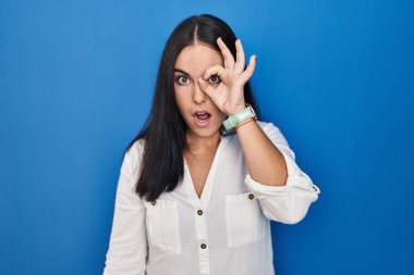 Mavi arka planda dikilen Latin kökenli genç bir kadın şaşırmış yüz ifadesiyle şaşırmış, parmaklarından bakan bir jest yapıyor. İnançsız ifade. 