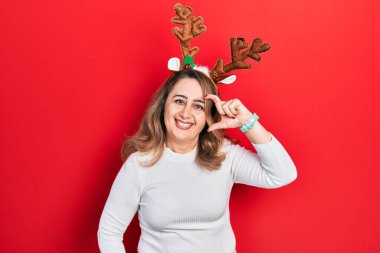 Orta yaşlı beyaz kadın şirin Noel geyiği boynuzları takıyor gülümsüyor ve kendine güvenen el hareketleriyle küçük beden işareti yapıyor parmaklarıyla ve kamerasıyla. ölçüm kavramı. 
