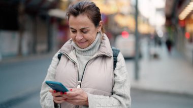 Orta yaşlı İspanyol kadın sokakta akıllı telefon kullanarak gülümsüyor.