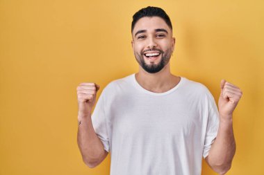 Sarı arka plan üzerine gündelik tişört giyen genç yakışıklı adam kollarını kaldırarak ve açık gözlerle başarının şaşkınlığını ve şaşkınlığını yaşıyor. Kazanan konsept. 