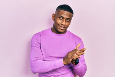 Genç siyah adam pembe bir süveter giyiyor, alkışlıyor, alkışlıyor, mutlu ve mutlu, gururla gülümsüyor. 