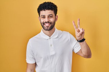 Sarı arka planda duran Arap adam, mutlu ve kendinden emin bir şekilde gülümserken iki numarayı işaret ediyor.. 