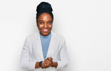 Genç Afro-Amerikan kadın, iş kıyafetleri içinde ellerini kavuşturup gülümseyerek gevşemiş ve neşeli gülümsüyor. Başarı ve iyimserlik 