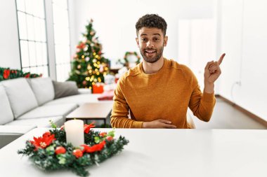 Arap genç adam noel ağacının yanında oturuyor, yüzünde kocaman bir gülümsemeyle, eli ve parmağı kameraya bakıyor.. 