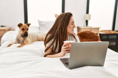 Genç İspanyol kadın dizüstü bilgisayar kullanıyor ve yatakta köpeklerle kahve içiyor.