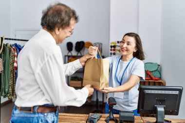 Orta yaşlı bir adam ve kadın, mağazada alışveriş çantası tutarak gülümsüyor.