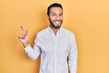 Sakallı, iş gömleği giyen, yüzünde kocaman bir gülümseme olan, eli ve parmağı kameraya bakan İspanyol bir adam.. 